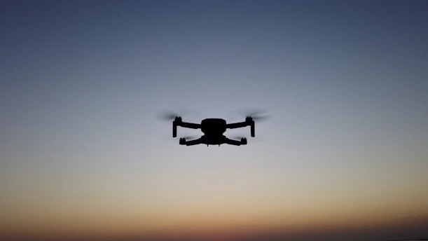 A sleek drone hovering against a bright baby blue sky with a hint of orange sunset in the background.