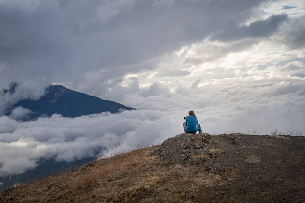 man in blue jacket sitting on brown rock mountain under white clouds during daytime