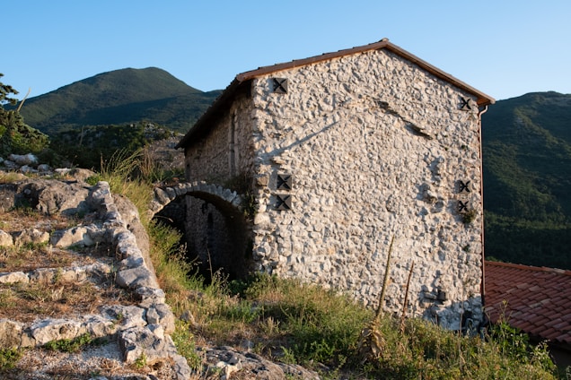 The image features a rustic stone building with a sloped tile roof, nestled in a mountainous landscape. Surrounding the structure are stone steps and sparse vegetation, illuminated by the warm glow of late afternoon sunlight. The mountains in the background provide a serene and secluded atmosphere.