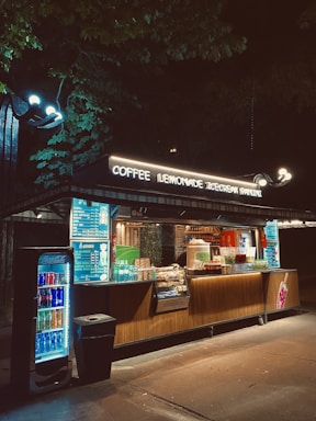 A well-lit food kiosk in a nighttime setting offers coffee, lemonade, ice cream, and panini. The kiosk has a wooden facade with a displayed menu and shelves stocked with various snacks and beverages. Bright lights illuminate the counter, and there's a fridge filled with assorted drinks visible. Surrounding trees darken the backdrop, enhancing the kiosk's illumination.