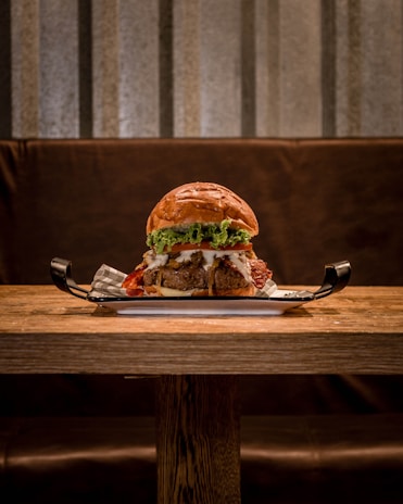 A large, appetizing burger with multiple layers including beef patties, lettuce, tomato, melted cheese, and bacon, placed on a flat serving tray atop a wooden table. The background features a muted, textured wall, creating a cozy dining ambiance.