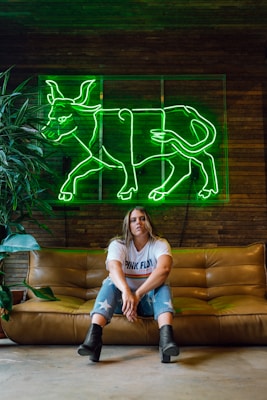 A person wearing a 'Pink Floyd' T-shirt and jeans with star designs sits on a brown leather couch. Above is a bright green neon sign in the shape of a bull. The setting has a rustic and modern feel, with wood paneling in the background and a potted plant to the side.