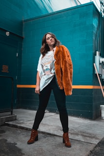 A person stands confidently in an industrial setting with a teal-colored wall in the background. They are wearing a graphic t-shirt, dark pants, brown heeled boots, and a rust-colored fur jacket draped over one shoulder.