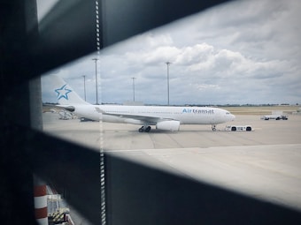 A large Air Transat airplane is parked on an airport tarmac, viewed through slatted blinds. The aircraft is white with a blue logo on the tail. There are several airport service vehicles nearby, including a pushback tug. Overhead, the sky is cloudy with a hint of blue visible.