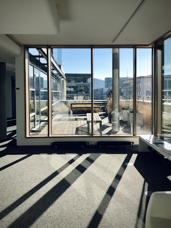 Sunlight pours through large glass windows onto a modern indoor space with a view of an exterior wooden deck. The scene includes contemporary architecture with flat roofs and angular lines, as well as planters containing tall grasses.