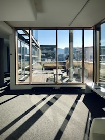 Sunlight pours through large glass windows onto a modern indoor space with a view of an exterior wooden deck. The scene includes contemporary architecture with flat roofs and angular lines, as well as planters containing tall grasses.