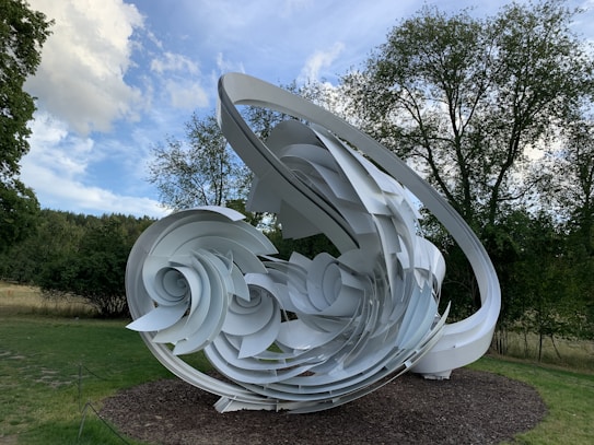 A large, abstract sculpture made of interlocking white spirals and curves is set outdoors on a grassy area, surrounded by trees and under a partly cloudy sky.