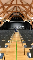 A spacious hall with a high wooden beam ceiling and rows of empty chairs facing a stage set up for a music performance with drums, guitars, and a keyboard.