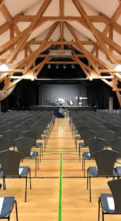 A spacious hall with a high wooden beam ceiling and rows of empty chairs facing a stage set up for a music performance with drums, guitars, and a keyboard.