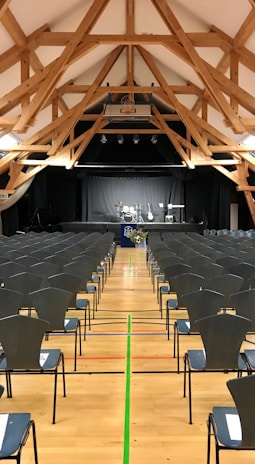 A spacious hall with a high wooden beam ceiling and rows of empty chairs facing a stage set up for a music performance with drums, guitars, and a keyboard.