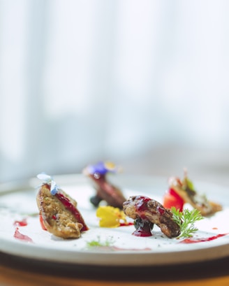 A gourmet dish is artfully presented on a plate with succulent pieces of meat garnished with colorful sauces and edible flowers. The dish includes vibrant splashes of red and yellow, complemented by green herbs, creating an elegant and sophisticated culinary presentation.