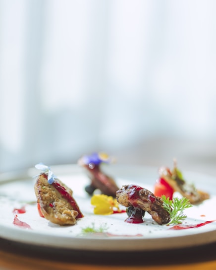 A gourmet dish is artfully presented on a plate with succulent pieces of meat garnished with colorful sauces and edible flowers. The dish includes vibrant splashes of red and yellow, complemented by green herbs, creating an elegant and sophisticated culinary presentation.