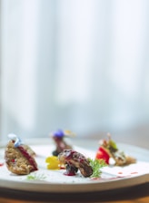A beautifully plated dish features several small, intricately arranged pieces of food on a white plate. Garnishes include edible flowers and sprigs of fresh herbs. The food is artfully presented with a drizzle of sauce and vibrant accents.