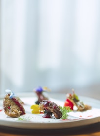 A beautifully plated dish features several small, intricately arranged pieces of food on a white plate. Garnishes include edible flowers and sprigs of fresh herbs. The food is artfully presented with a drizzle of sauce and vibrant accents.