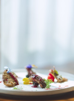 A beautifully plated dish features several small, intricately arranged pieces of food on a white plate. Garnishes include edible flowers and sprigs of fresh herbs. The food is artfully presented with a drizzle of sauce and vibrant accents.