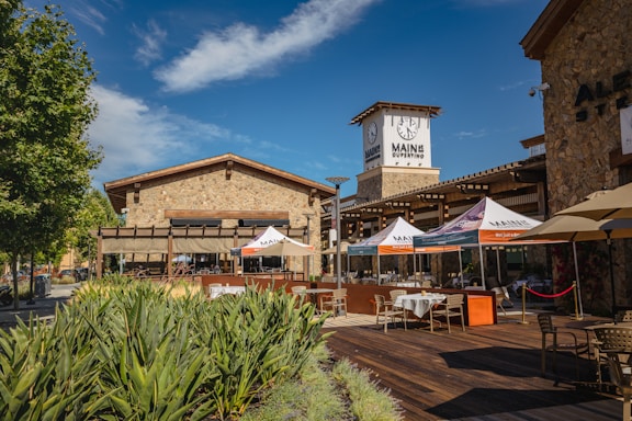 A rustic outdoor dining area is situated in front of a stone building with a large clock tower. Several tables are set up, covered with umbrellas displaying the name 'Main'. A clear blue sky and a row of green plants add to the vibrant atmosphere.