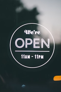 A circular sign with a black background displaying white text that reads 'We’re OPEN' with operating hours from 11 AM to 11 PM.