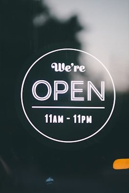 A circular sign with a black background displaying white text that reads 'We’re OPEN' with operating hours from 11 AM to 11 PM.