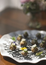 An artistic arrangement of dumplings is presented on a white plate, decorated with black twig-like garnishes and colorful flowers. Each dumpling is topped with a clear cube and a thin green garnish, offering a visually appealing gourmet experience.