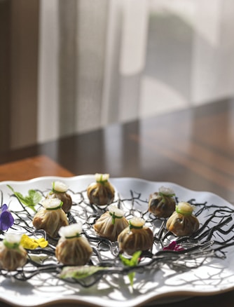 A white, scalloped-edged plate is arranged with intricately designed dumplings, garnished with small green herb leaves and floral elements. The plate sits on a wooden table bathed in natural light, casting soft shadows.