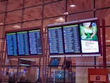 Four electronic display boards are mounted in an airport terminal, showing flight departure information. Two screens show domestic departures, and two show international departures. A separate screen displays an advertisement promoting environmental care with a green theme. The airport interior has a modern design, and the ceiling features geometric patterns with ambient lighting.