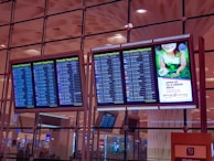 Four electronic display boards are mounted in an airport terminal, showing flight departure information. Two screens show domestic departures, and two show international departures. A separate screen displays an advertisement promoting environmental care with a green theme. The airport interior has a modern design, and the ceiling features geometric patterns with ambient lighting.