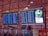 Four electronic display boards are mounted in an airport terminal, showing flight departure information. Two screens show domestic departures, and two show international departures. A separate screen displays an advertisement promoting environmental care with a green theme. The airport interior has a modern design, and the ceiling features geometric patterns with ambient lighting.