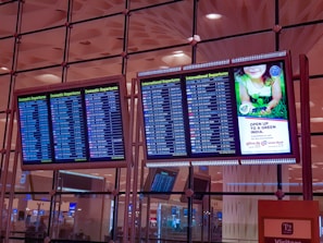 Four electronic display boards are mounted in an airport terminal, showing flight departure information. Two screens show domestic departures, and two show international departures. A separate screen displays an advertisement promoting environmental care with a green theme. The airport interior has a modern design, and the ceiling features geometric patterns with ambient lighting.
