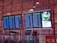 Four electronic display boards are mounted in an airport terminal, showing flight departure information. Two screens show domestic departures, and two show international departures. A separate screen displays an advertisement promoting environmental care with a green theme. The airport interior has a modern design, and the ceiling features geometric patterns with ambient lighting.