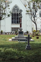 A cemetery featuring two stone crosses with engraved text is situated on a grass lawn in front of a large, white gothic-style church. The church has pointed arch windows with intricate designs, and there are two trees with sparse foliage on either side.