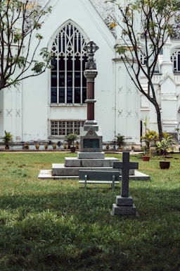A cemetery featuring two stone crosses with engraved text is situated on a grass lawn in front of a large, white gothic-style church. The church has pointed arch windows with intricate designs, and there are two trees with sparse foliage on either side.