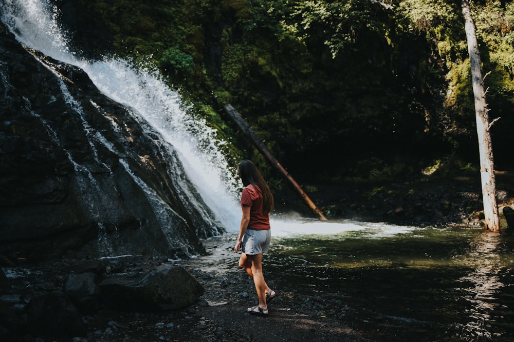 woman in blue shirt and blue denim shorts standing on black soil near waterfalls during daytime