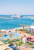 A vibrant waterfront scene depicting a beach resort with a water park in the foreground. The water park features slides, orange lounge chairs, and umbrellas. Beyond the park, a beach and a harbor with a large cruise ship are visible, surrounded by a clear blue sky.