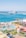 A vibrant waterfront scene depicting a beach resort with a water park in the foreground. The water park features slides, orange lounge chairs, and umbrellas. Beyond the park, a beach and a harbor with a large cruise ship are visible, surrounded by a clear blue sky.