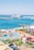 A vibrant waterfront scene depicting a beach resort with a water park in the foreground. The water park features slides, orange lounge chairs, and umbrellas. Beyond the park, a beach and a harbor with a large cruise ship are visible, surrounded by a clear blue sky.