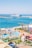 A vibrant waterfront scene depicting a beach resort with a water park in the foreground. The water park features slides, orange lounge chairs, and umbrellas. Beyond the park, a beach and a harbor with a large cruise ship are visible, surrounded by a clear blue sky.