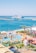 A vibrant waterfront scene depicting a beach resort with a water park in the foreground. The water park features slides, orange lounge chairs, and umbrellas. Beyond the park, a beach and a harbor with a large cruise ship are visible, surrounded by a clear blue sky.