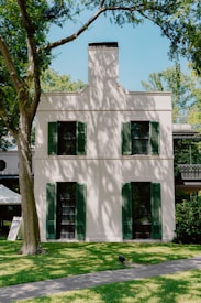 A two-story building with light-colored walls and dark green shutters on the windows, surrounded by lush green trees and neatly manicured grass. A pathway leads up to the building, and dappled sunlight creates patterns across the facade.