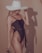 A woman wearing a dark one-piece swimsuit and a large, wide-brimmed white hat. Her long blonde hair is visible, and she is slightly turned to the side, holding sunglasses in one hand. The background is a plain beige color, and her shadow is cast on the wall.
