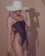 A woman wearing a dark one-piece swimsuit and a large, wide-brimmed white hat. Her long blonde hair is visible, and she is slightly turned to the side, holding sunglasses in one hand. The background is a plain beige color, and her shadow is cast on the wall.