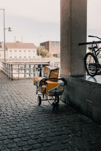 An urban scene featuring a cobblestone walkway under a covered area. A cart with yellow and silver postal bags is parked next to a railing. A bicycle is partially visible on the right side of the image. In the background, there are buildings, a fence, and an outdoor area with closed umbrellas.