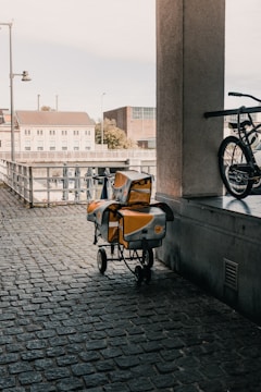An urban scene featuring a cobblestone walkway under a covered area. A cart with yellow and silver postal bags is parked next to a railing. A bicycle is partially visible on the right side of the image. In the background, there are buildings, a fence, and an outdoor area with closed umbrellas.
