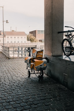 An urban scene featuring a cobblestone walkway under a covered area. A cart with yellow and silver postal bags is parked next to a railing. A bicycle is partially visible on the right side of the image. In the background, there are buildings, a fence, and an outdoor area with closed umbrellas.