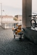 An urban scene featuring a cobblestone walkway under a covered area. A cart with yellow and silver postal bags is parked next to a railing. A bicycle is partially visible on the right side of the image. In the background, there are buildings, a fence, and an outdoor area with closed umbrellas.