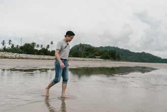 A smiling traveler standing barefoot on the warm sand, with lush green hills in the background.