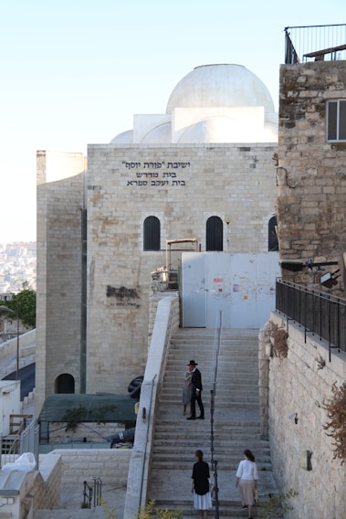A stone building with Hebrew text on it, featuring arched windows and a dome on the roof. There are stairs leading up to the structure with several people, some dressed in traditional clothing, walking on them. The surroundings include a stone wall and a small paved path.