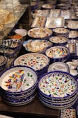 A collection of colorful, intricately designed ceramic plates and bowls, featuring vibrant patterns and motifs. The dishes are neatly stacked and displayed on a surface, with some glassware in the background.