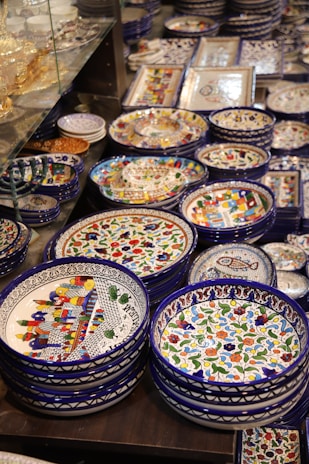 A collection of colorful, intricately designed ceramic plates and bowls, featuring vibrant patterns and motifs. The dishes are neatly stacked and displayed on a surface, with some glassware in the background.