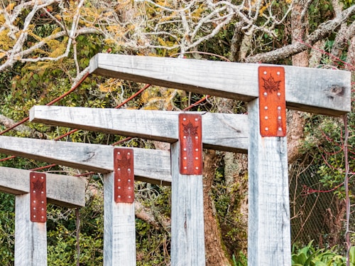 Several wooden beams with red metal brackets are arranged in a row, flanked by a densely wooded area. The beams are weathered, and the metal brackets are decorated with a leaf pattern. The trees in the background have elaborate branches and a mix of green and orange foliage.