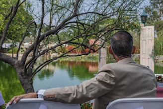 A person in a beige suit is seated outdoors, overlooking a serene pond surrounded by lush greenery and trees. The scene includes an open doorway structure near the water's edge, with a lamp post visible in the background. The person's arm rests on the back of a white chair, creating a relaxed atmosphere.